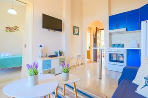 A modern living room with a sofa, a dining table with two potted plants, a kitchen with blue cabinets and a white refrigerator, and a flat-screen TV mounted on the wall. - Evora in Ionian Islands (Ionian Islands)
