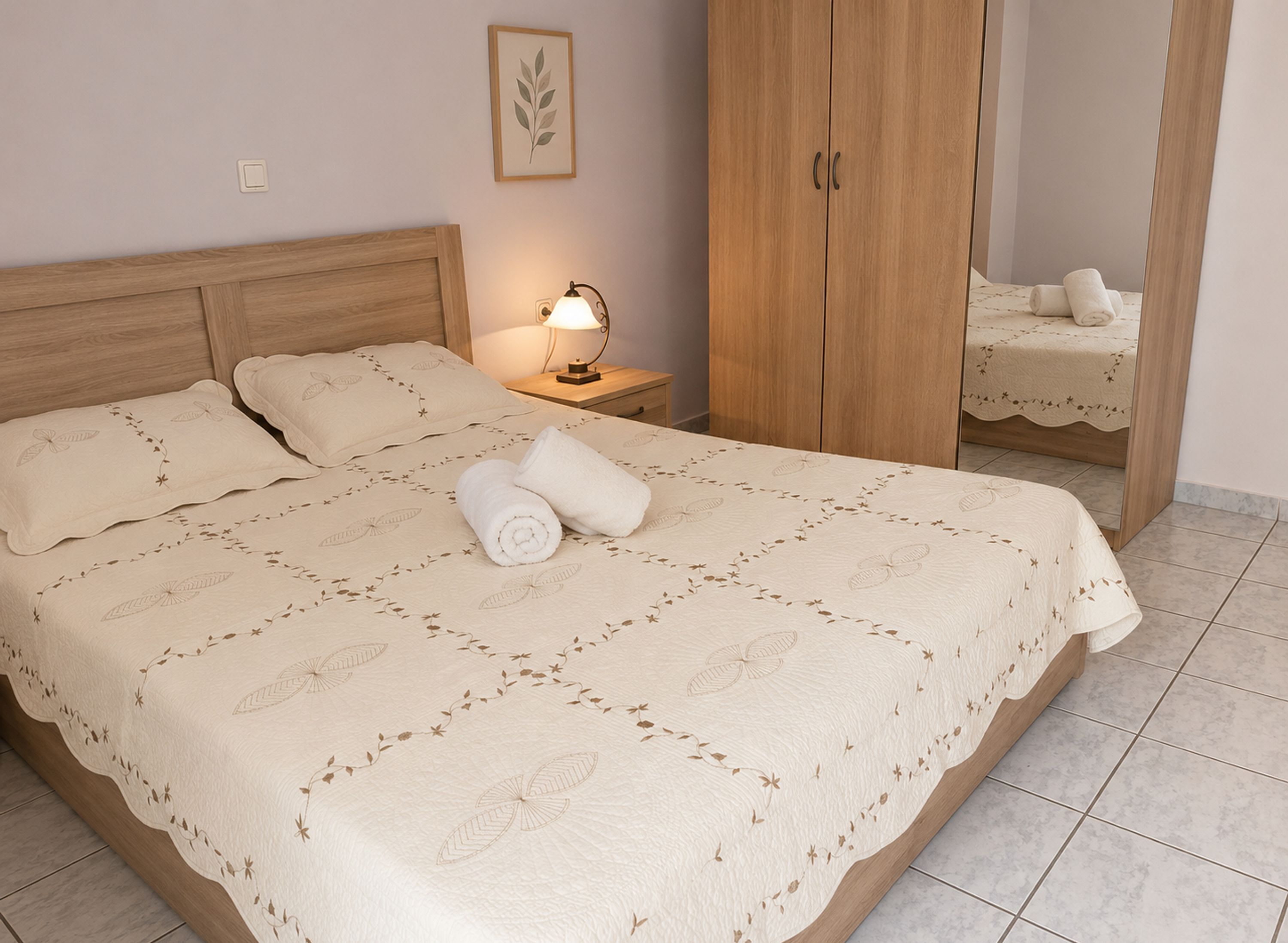 A neatly made bed with a floral-patterned bedspread, a nightstand with a lamp, a wooden wardrobe, and a mirror reflecting the bed.