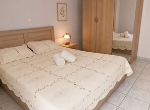 Ein ordentlich bezogenes Bett mit floralem Überwurf, ein Nachttisch mit Lampe, ein Holzschrank und ein Spiegel, der das Bett reflektiert.