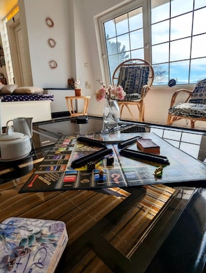 A room with a large window, a glass-top table with board games, wicker chairs, and a vase with flowers.