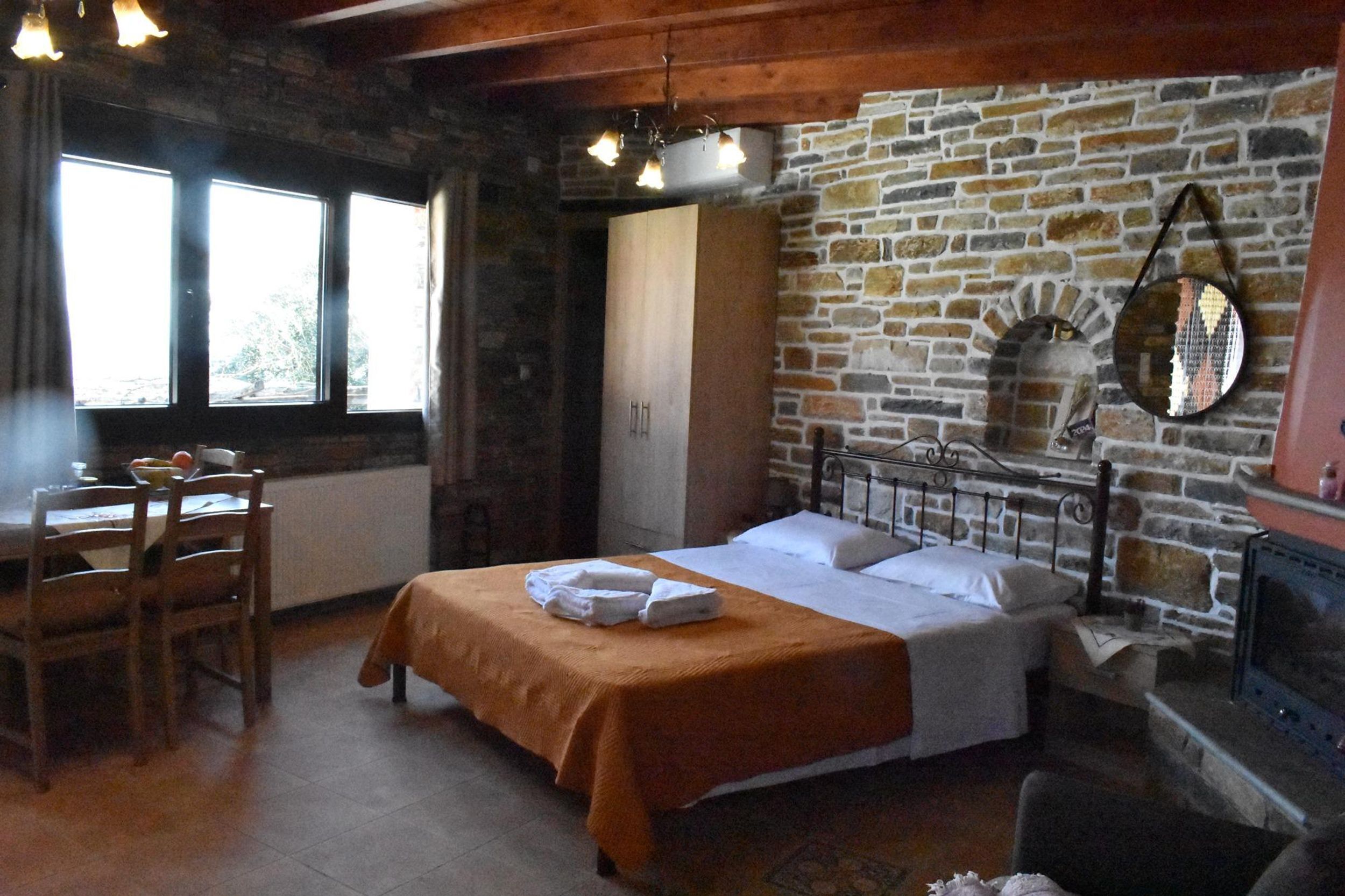 A room with a bed, a wooden table with chairs, a stone wall, and a fireplace.