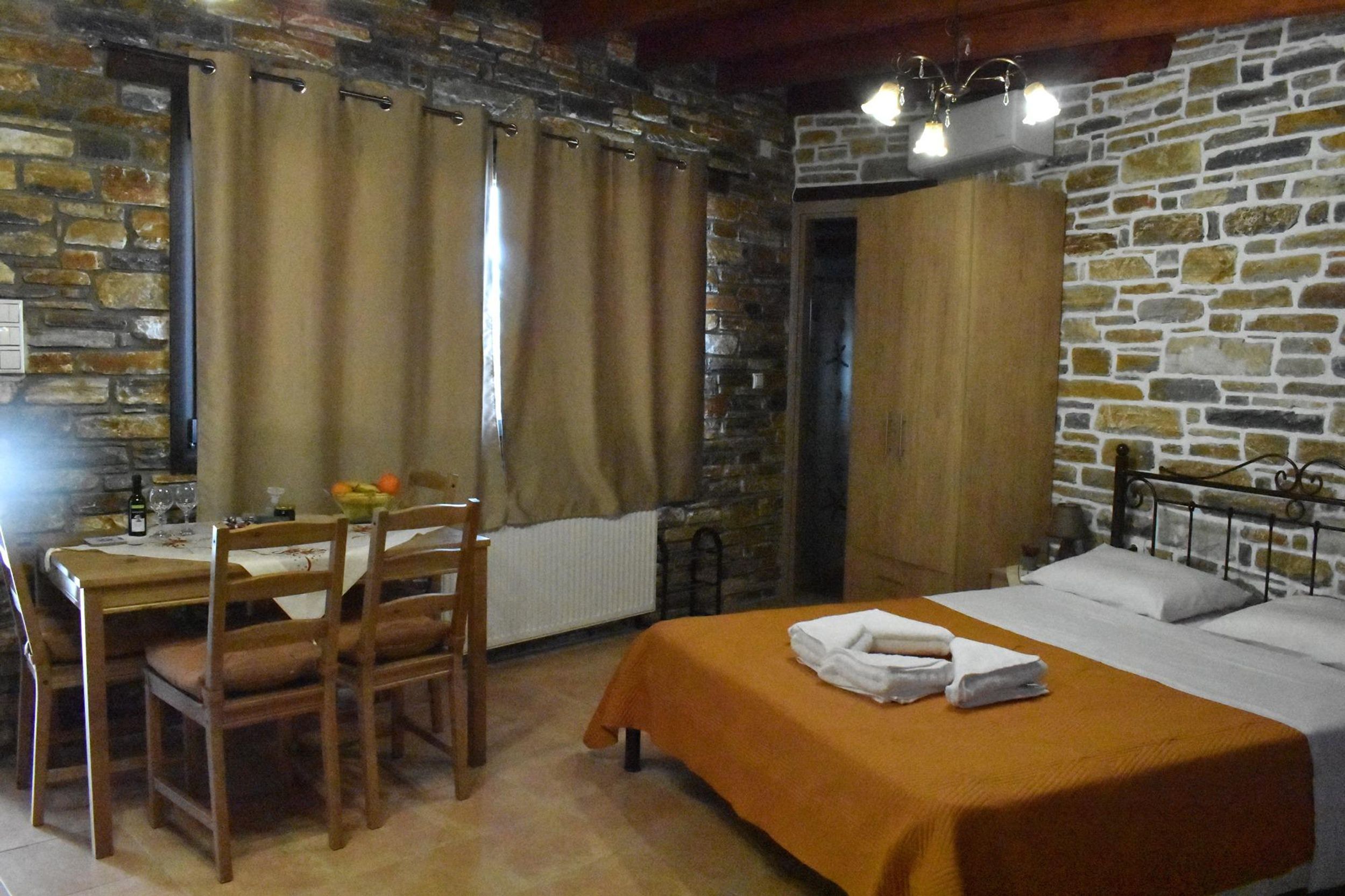 Ein Zimmer mit Steinwand, einem Bett mit orangefarbener Bettdecke, einem Holztisch mit Stühlen und einem Fenster mit Vorhängen.