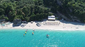 Vista aérea de una playa con aguas turquesas cristalinas, un promontorio rocoso y un edificio con techo blanco.