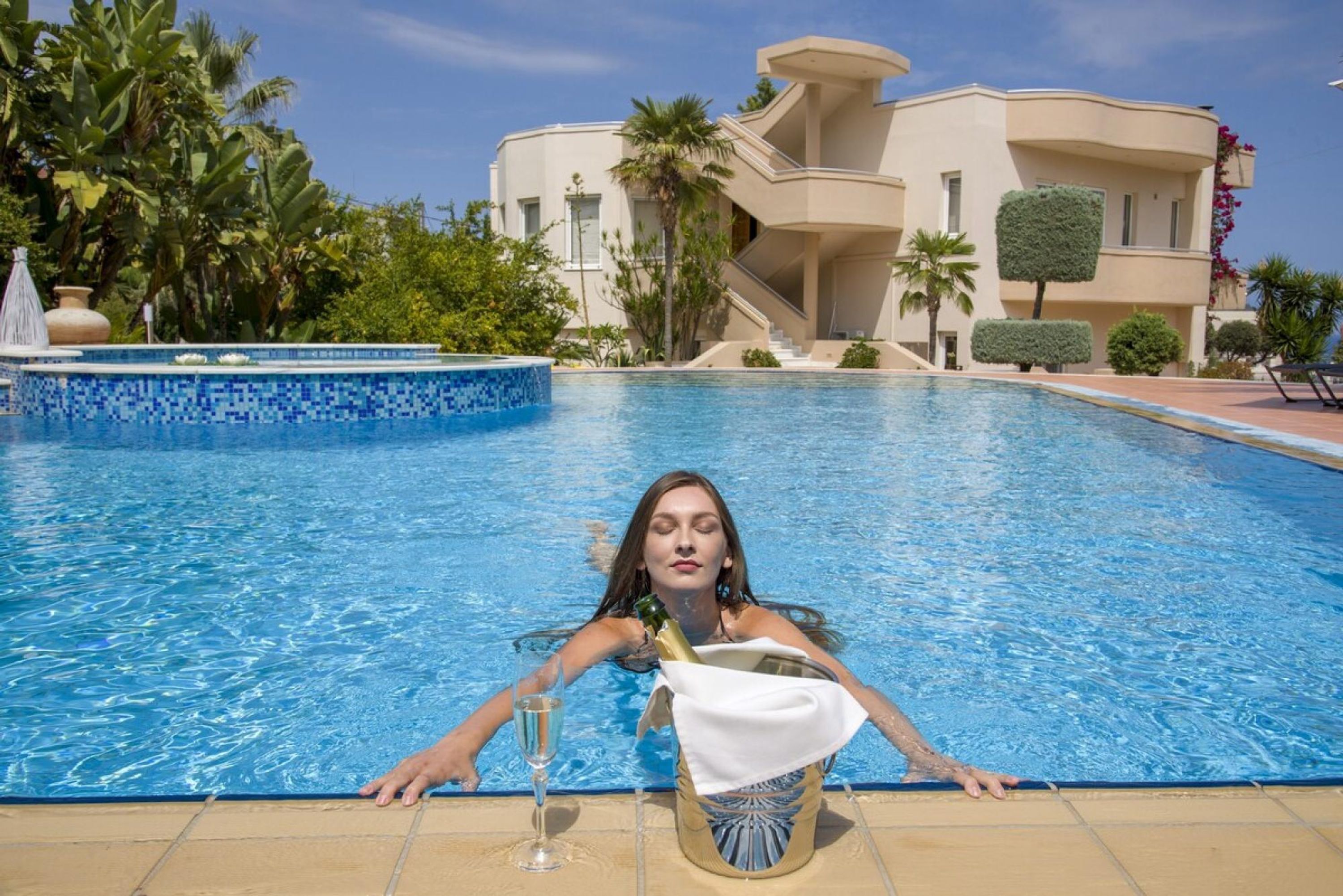 A pool area with a clear blue pool, a chair, and a bottle of champagne.