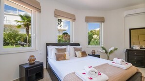 Ein Schlafzimmer mit einem großen Bett, zwei Nachttischen, einer Kommode und einem Fenster mit Blick auf Grünflächen und ein Gebäude.