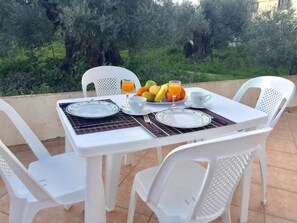 Et bord dækket til morgenmad med tallerkener, bestik og en skål med frugt på en terrasse med hvide stole og udsigt til oliventræer.