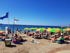 A beach with multiple colorful umbrellas and sun loungers, a flag, and a signboard. - Mirsini's Apartment in Chania center (Chania)