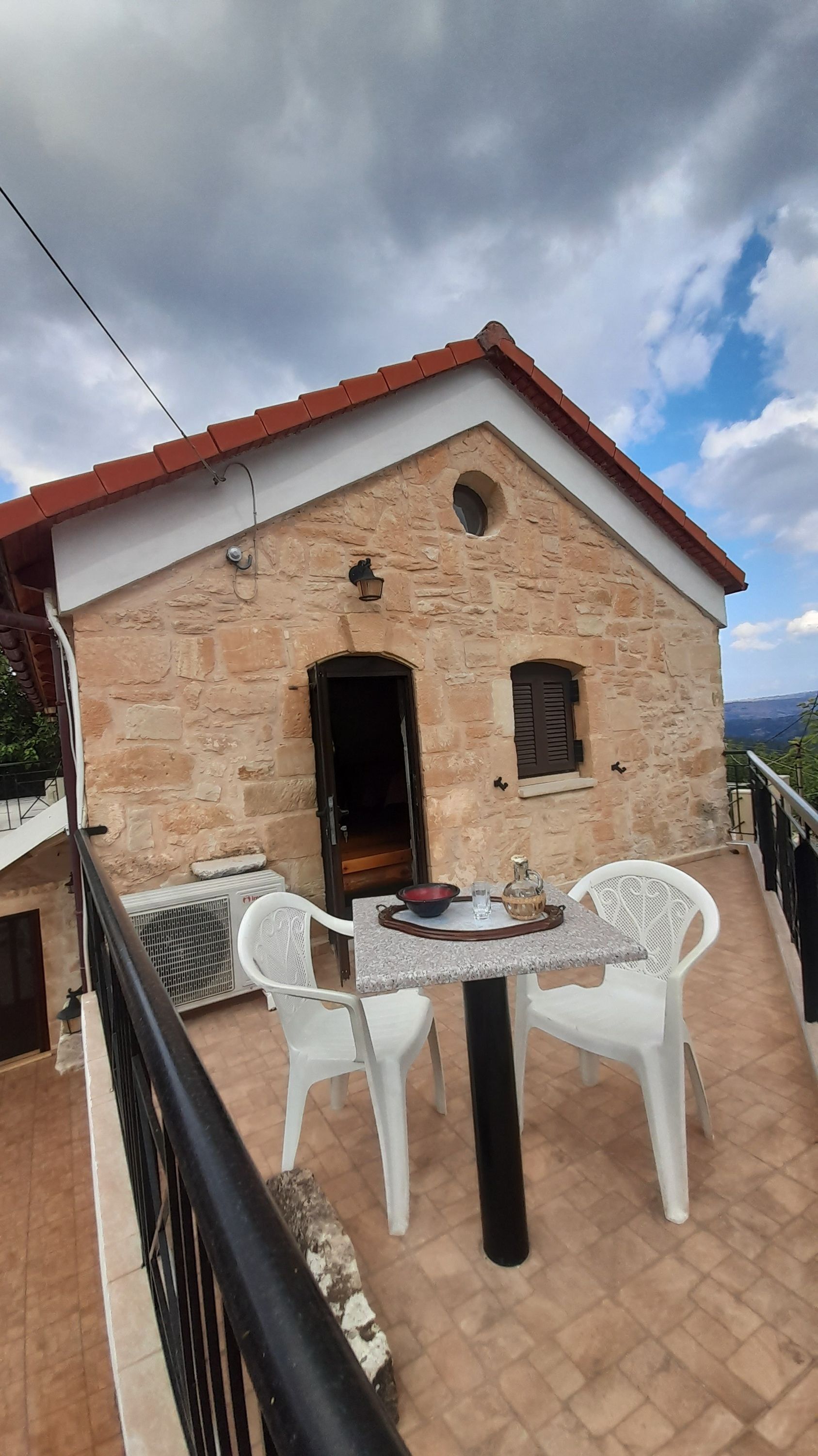 A stone building with a red roof, a small balcony with white plastic chairs, and a table with a bowl and a glass.
