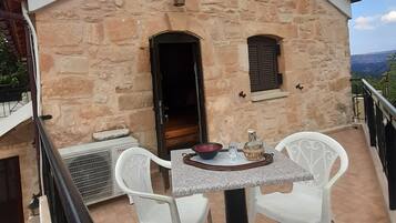 A stone building with a red roof, a small balcony with white plastic chairs, and a table with a bowl and a glass.