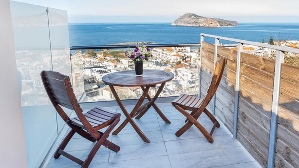 Un balcon avec deux chaises en bois et une petite table ronde, d’où l’on peut admirer une ville côtière et la mer.
