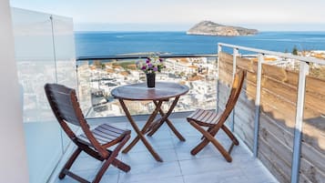 Un balcon avec deux chaises en bois et une petite table ronde, d’où l’on peut admirer une ville côtière et la mer.