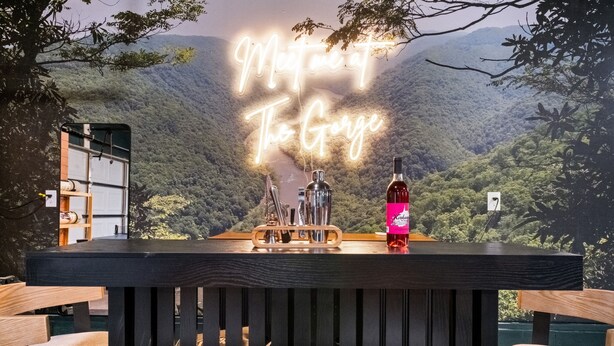 Eine Bar mit einem Neonlichtschild mit der Aufschrift "Meet me at The Gorge", Holzsesseln und einer Weinflasche auf einem Tisch mit Blick auf ein bewaldetes Tal.