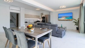 A modern living space with a dining area, kitchen, and a large window with a view of the sea. - Villa Erato – Agios  Onoufrios (Kounoupidiana)