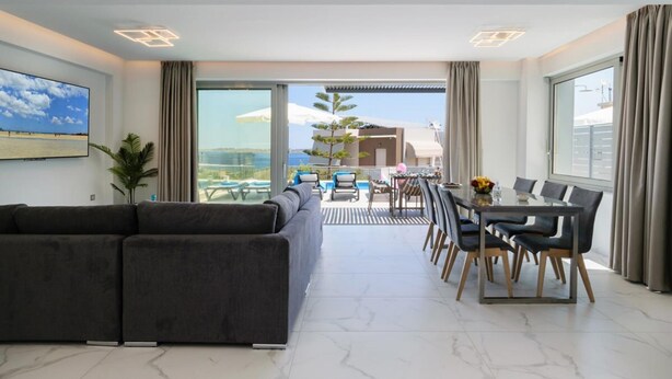 A modern living room with a sofa, dining table, and chairs, a balcony with a pool view, and large windows. - Villa Erato – Agios  Onoufrios (Kounoupidiana)
