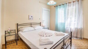 Ein ordentlich bezogenes Bett mit weißen Bettwäsche und Handtüchern, ein Nachttisch mit Blumen in einer Vase, ein Fenster mit durchsichtigen Vorhängen und eine Tür im Hintergrund.