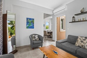 Ein modernes Wohnzimmer mit einem grauen Sofa, einem Holztisch und einem grauen Sessel. An der Wand hängt ein Gemälde und das Fenster bietet Blick auf Grünflächen.