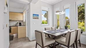 A modern kitchen with wooden cabinets, a dining area with a dark wooden table and beige upholstered chairs, and large windows offering a view of greenery. - Villa Blue Dreams in Maleme (Maleme)