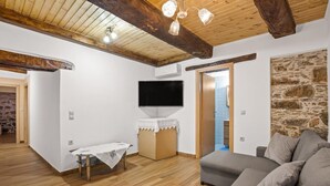 A cozy living room with a wooden ceiling, a stone wall, a grey sofa, and a television mounted on the wall. - Maria's House with View in Kambos (Kampos)