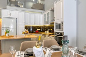 A modern kitchen with white cabinets, a marble countertop, and a dining area set for two with a bottle of champagne. - Villa Hope in Chania (Mournies)