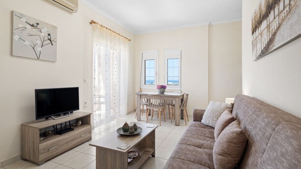 Een woonkamer met een flatscreen televisie, een houten salontafel, een bank, een eethoek met een tafel en stoelen, en een raam met gordijnen.