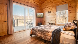 A wooden cabin with a bed, a television, and a sliding glass door leading to an outdoor deck. - Brand New Luxury Cabin On 2.7 Acres | Fire Pit (Berkeley Springs)