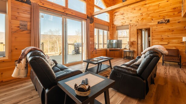 A log cabin interior with a fireplace, leather seating, a television, and large windows. - Brand New Luxury Cabin On 2.7 Acres | Fire Pit (Berkeley Springs)