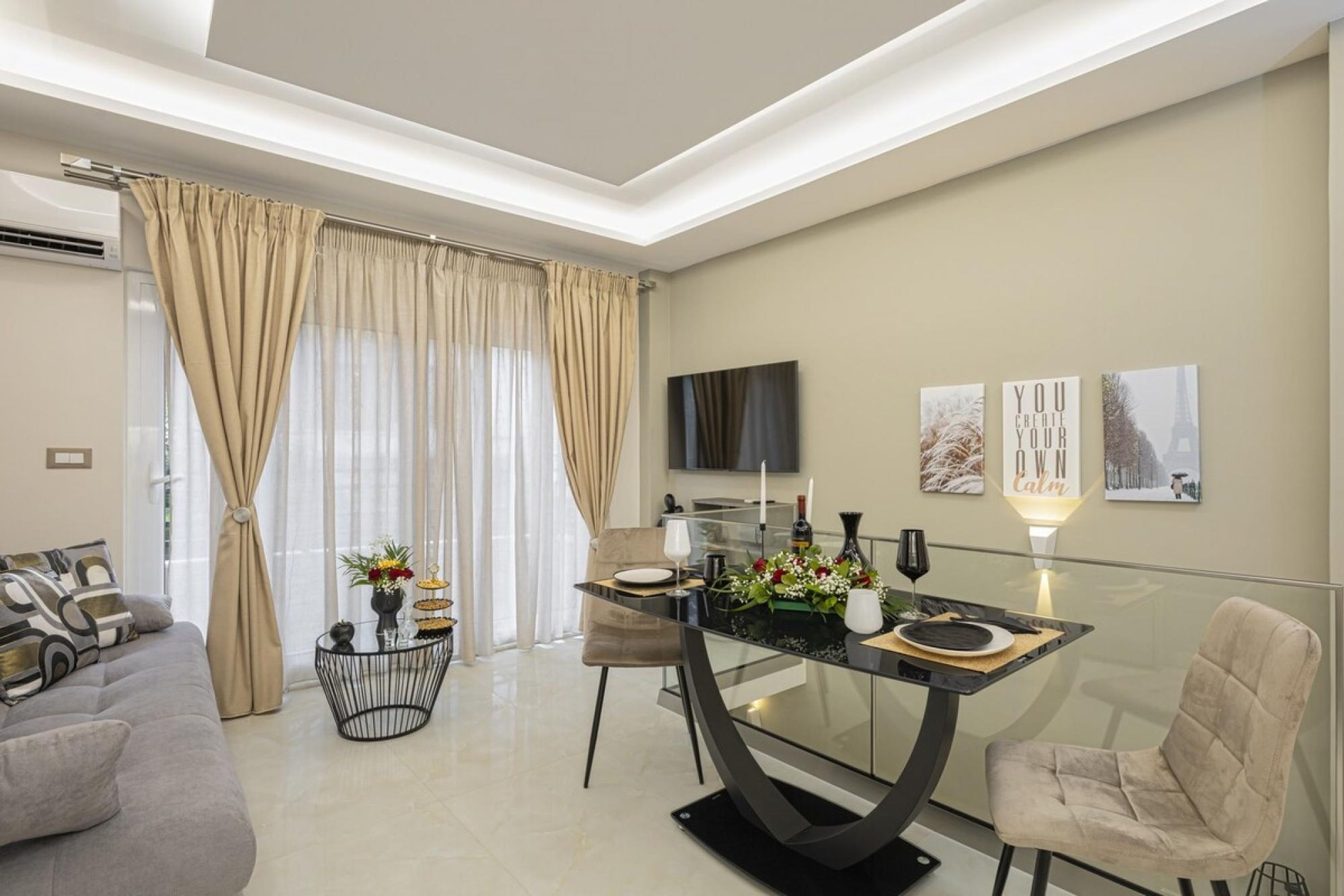 Una sala moderna con una mesa de comedor de vidrio, cortinas color beige, un sofá y un televisor de pantalla plana montado en la pared.