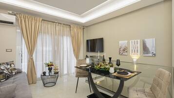 Una sala moderna con una mesa de comedor de vidrio, cortinas color beige, un sofá y un televisor de pantalla plana montado en la pared.