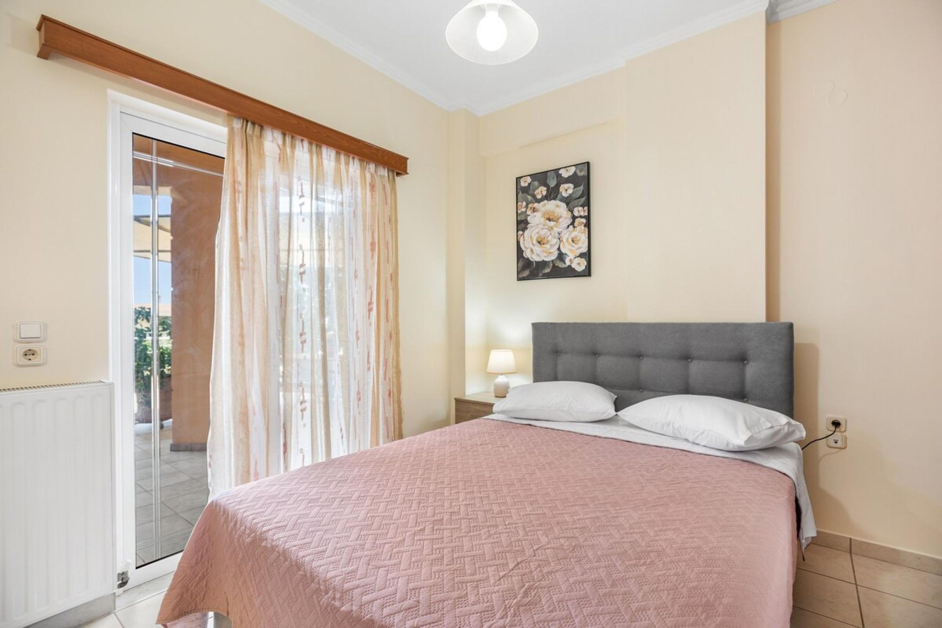 Een slaapkamer met een bed, nachtkastje, lamp, raam met gordijnen en een schilderij aan de muur.