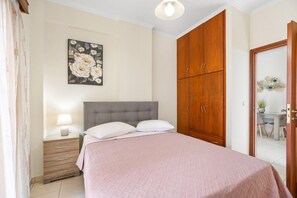 A bedroom with a bed, a wooden wardrobe, a nightstand with a lamp, and a floral painting on the wall. - Sofie's Garden View Apartment (Daratsos)