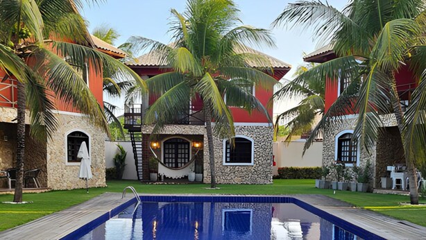 A pool area with a clear blue pool, surrounded by palm trees and a stone building with red and white walls. - Chalé De Canoa (Aracati)