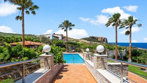 A pool area with stone railings, palm trees, and a view of the sea. - Kambos Horizon Villa (Kampos)