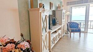 Un salon spacieux doté d’une grande fenêtre donnant sur la mer, d’un meuble de rangement en bois et d’un fauteuil bleu à motifs.