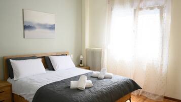 A neatly made bed with a dark bedspread and white pillows, a bedside lamp, and a window with sheer curtains.