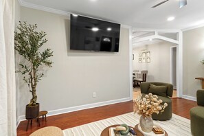 A modern living room with a flat-screen TV mounted on the wall, a large potted plant, a green sofa, and a coffee table with decorative items.