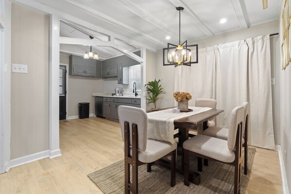 A dining area with a wooden table, four chairs, a chandelier, and a view into a kitchen.