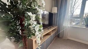 A living room with a large plant, a flat-screen TV on a wooden stand, a lamp, and a window with curtains. - Central Horsham Flat | Free Parking (Horsham)