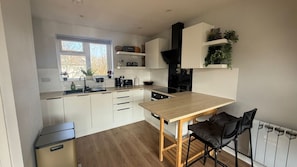 Une cuisine moderne avec des meubles blancs, une table en bois et un plan de travail noir. Une fenêtre laisse entrer la lumière naturelle, et un radiateur est visible sur la droite.