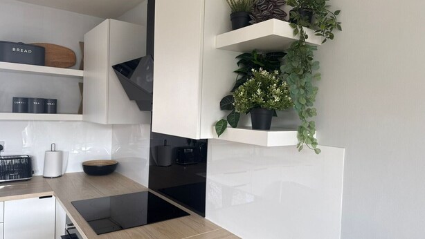 A modern kitchen with white cabinetry, a black countertop, and a built-in stove. There are shelves with potted plants and a bread box on the upper shelf. - Central Horsham Flat | Free Parking (Horsham)