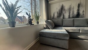 A modern living room with a grey sectional sofa, a small ottoman, and a large window with a view of houses and trees. - Central Horsham Flat | Free Parking (Horsham)
