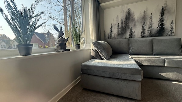 Un salon moderne avec un canapé d’angle gris, un petit pouf et une grande fenêtre donnant sur des maisons et des arbres.