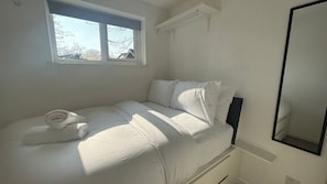 Un lit bien fait, avec du linge de lit blanc et des oreillers, une fenêtre donnant sur des arbres et des immeubles, et un miroir accroché au mur.