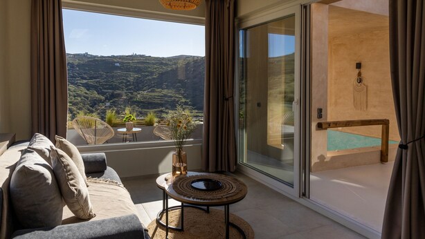 A modern living room with a large window, a sofa, a small table, and a view of a pool and hills. - Gaea Dream Villas Complex (Tinos ,Loutra)