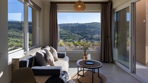 A modern living room with a sofa, a small table, and a view of a mountainous landscape through large windows. - The Pyr Element Of Gaea (Loutra)