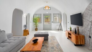 Een moderne woonkamer met een grijze bank, een houten salontafel en een grijze poef. Er is een houten tv-meubel met een flatscreen televisie die aan een stenen muur is bevestigd. Een groot raam met uitzicht op groen is zichtbaar.