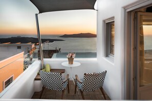 En balkon med bord og stole, der ud til et kystlandskab i solnedgang.