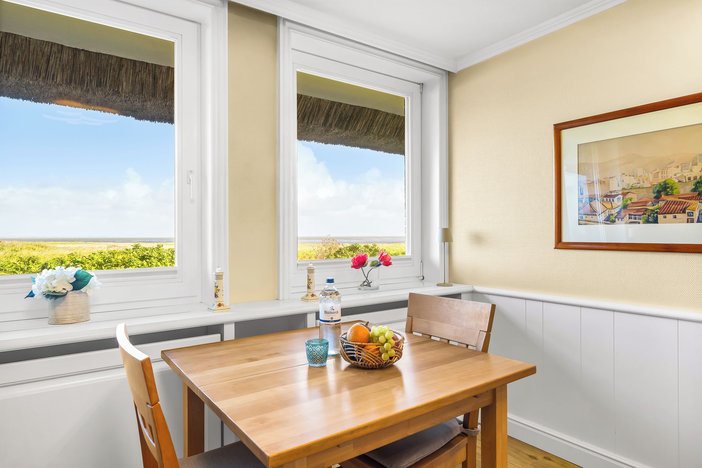 A dining area with a table, chairs, a vase with flowers, a bottle, and a basket of fruit.