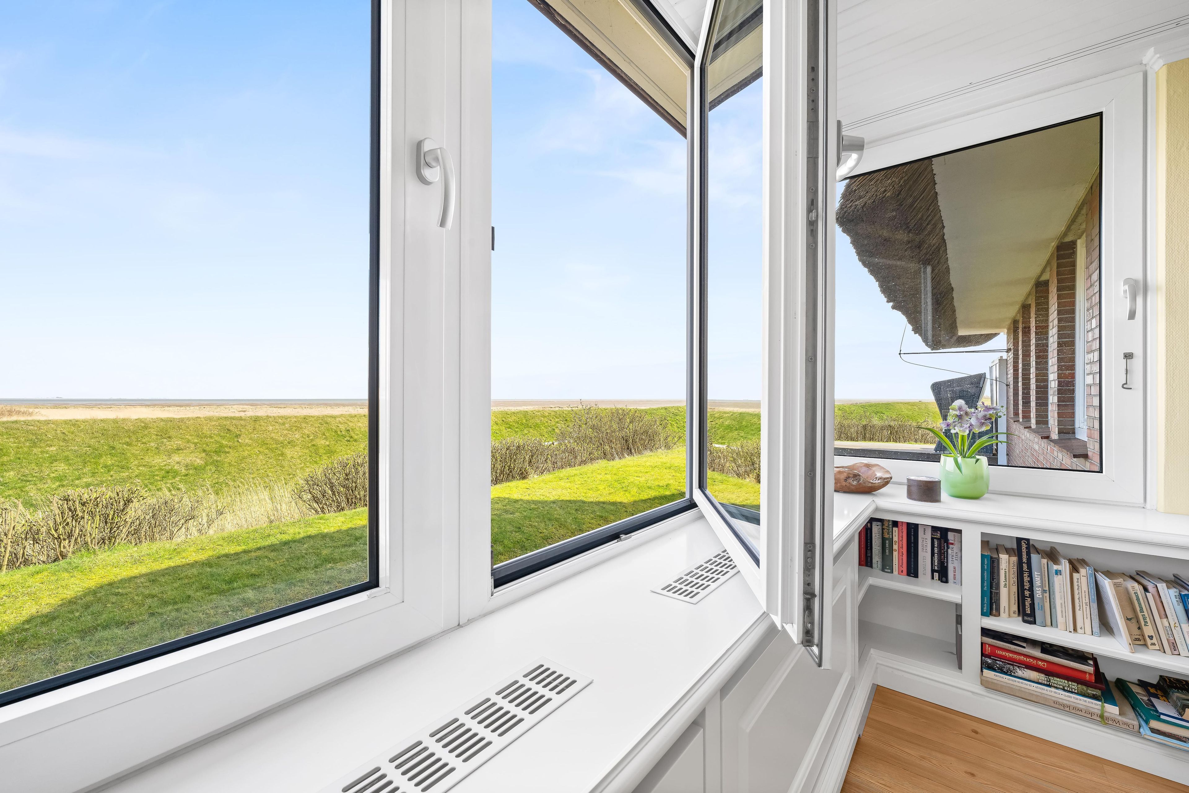 A room with large windows offering a view of a grassy field. There is a bookshelf with books and a small vase with flowers.