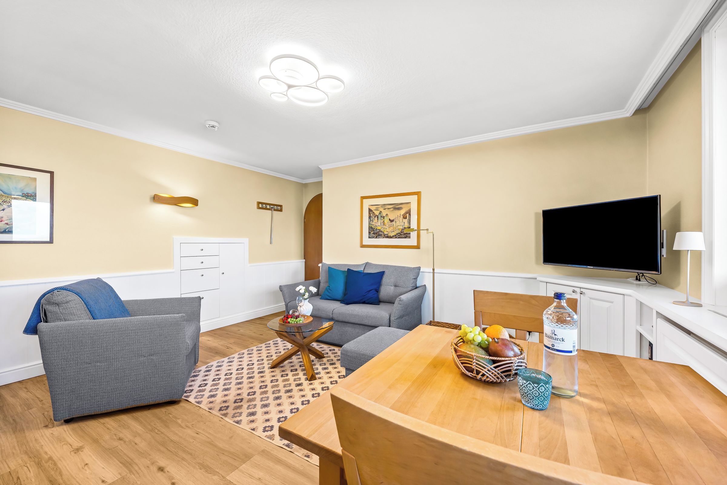 A modern living room with a grey sofa, a wooden dining table with fruit, a flat-screen TV, and a ceiling light fixture.