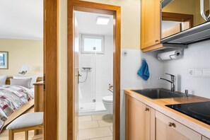 A compact hotel room with a wooden door leading to a shower, a bed with a quilt, a sink with a faucet, and a small kitchen area with cabinets. - Watt Apartment, right on Rantum Bay on Sylt (Sylt)